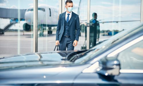 Businessman with a bag in hand standing near the glass door of an airport and looking at the car in front of him. Medical mask on his face